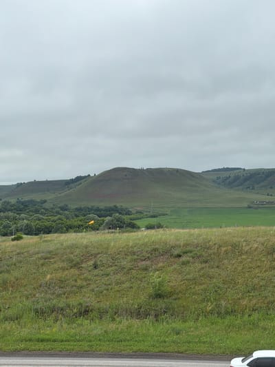 The image depicts a rural landscape under an overcast sky. In the foreground, a road with white lines is visible, and a white car is parked on the right side. The middle ground features a grassy embankment with scattered wildflowers and shrubs. Further back, rolling green hills dominate the scene, with trees dotting the slopes and a distant village with a few houses visible on the right. A yellow windsock is planted in the grass on the left side, and a few small figures can be seen on the crest of the largest hill. The overall environment suggests a temperate climate with lush vegetation. The location is specified as Kendektamak, Russia.