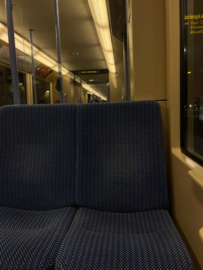 A textured, two-section blue and yellow seat is in the foreground. Beyond it, a window reveals a train platform at Felix-Littenkruke station. The interior of the train includes beige paneling, overhead lighting, and handrails. Window text displays destinations. The view outside showcases the station's architecture and signage.