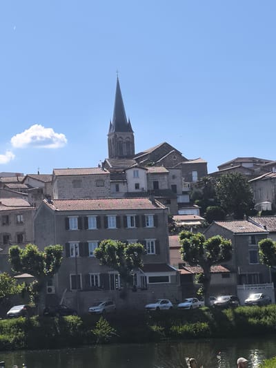 The photograph captures a view of Saint-Pierre-de-Bœuf, France, showcasing the town's architecture and waterfront. Buildings with red-tiled roofs and stone facades are clustered together, rising on a hillside. A prominent church with a tall, dark spire dominates the skyline. Trees with rounded crowns line the waterfront, partially obscuring a row of parked cars. A body of water reflects the scene. In the foreground, the heads of people suggest their presence on the waterfront. A few wispy clouds dot the blue sky.
