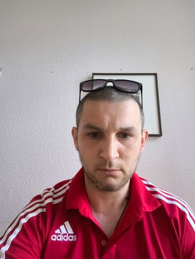 A medium shot photograph shows a man with short hair and a beard, wearing sunglasses on his head, and a red Adidas shirt with white stripes. He is looking at the camera with a neutral expression. Behind him, a framed picture is hanging on a textured, light-colored wall. The image appears to be taken indoors, possibly in a room in Greifswald, Germany, as suggested by the context.