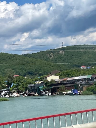 A wide-angle, eye-level shot captures a serene waterscape in Lermontovo, Russia, under a partly cloudy sky. A turquoise body of water dominates the foreground, with a red and white railing of a bridge or embankment running along the bottom edge of the frame. Along the opposite shore, a cluster of buildings and boats are visible. These structures appear to be part of a marina or a waterfront settlement, with several boats docked. Beyond the buildings, a lush, green, tree-covered hillside rises, forming a natural backdrop. Two tall, thin towers are visible on the crest of the hill. The overall scene suggests a tranquil, picturesque view of a coastal town or resort area.