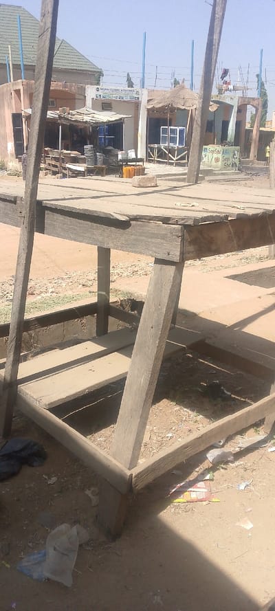 The image depicts an outdoor commercial scene in Danbare, Nigeria, under a clear, bright sky. In the foreground, an empty, weathered wooden market stall or table occupies the lower half of the frame. It is constructed from rough-hewn, dark planks forming a flat top and a lower frame, supported by sturdy vertical and angled beams. The ground immediately around the stall is dry, light-brown earth, scattered with debris including black and white plastic bags and other refuse. Pronounced shadows from the wooden structure are visible on the ground.

In the midground, beyond the foreground structure, a street or open area extends towards a row of single and two-story commercial buildings. On the left, a light-colored building, possibly beige or pale pink, features several dark windows and an open storefront area under a corrugated metal awning. Inside, various items, including multiple metal basins, are visible. Above this building, a horizontal sign reads "MISAN GLOBAL VENTURES" with additional smaller text and product images. Adjacent to the "MISAN GLOBAL VENTURES" building, a stall with a thatched roof canopy displays two white window frames on a wooden stand. To the right of this, a low concrete wall or structure is painted with text including "BKSnyaSpot ABG" and "Sarya Spot." Several yellow plastic jerry cans and a light-colored stone block are on the ground in this midground area.

In the background, additional buildings are visible, some with green corrugated metal roofs, others with flat roofs showing construction elements. Several thin, light blue vertical poles or pillars extend upwards from behind the buildings into the clear blue sky. Indistinct human figures are visible near the shops and further in the background; their actions are not discernible. Sparse green vegetation is visible in the distant background.