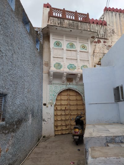 A wide shot captures a narrow, dusty alleyway in Jodhpur, India, flanked by textured, blue-painted walls on the left and light blue-painted structures on the right. The focal point is a large, arched, yellow metal door with ornate dark metal detailing, set within a cream-colored building adorned with green decorative accents around the arch and on the balcony above. The balcony, featuring white shutters and decorative greenish-blue inlays, is topped by a red crenellated parapet. A parked yellow scooter with a black seat and a license plate that reads "RJ19S R2083" sits to the right of the door. The ground is a mixture of dirt and paving stones. Overhead, a cloudy sky is visible.
