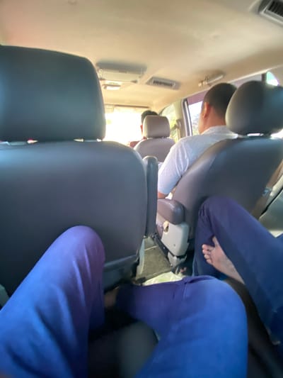 The image shows the interior of a car, likely an SUV, with four people visible. The perspective is from the rear passenger seats, looking forward. In the foreground, the legs of two individuals wearing dark blue pants are prominent. One person's bare feet are visible.
Further forward, the back of a driver's seat is seen, followed by the heads and shoulders of two men in the middle row. The man on the right is wearing a white patterned shirt and looking forward. The man to his left is mostly obscured by the headrest, but appears to be wearing a red shirt.
The car interior is illuminated by daylight, suggesting it is daytime. The dashboard and air vents are partially visible. The environment outside the car is out of focus but appears to be a bright, possibly sunny, outdoor scene with some greenery. The presence of a car interior with passengers suggests a mode of transportation within a populated area. The mention of Aluepiet, Indonesia, implies the location is within this specific city.