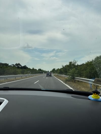 The image provides a forward-facing view from inside a moving vehicle, looking out onto a highway, reportedly in Zagore, Bulgaria. The car's dashboard is visible in the foreground, primarily on the right side of the frame.
A distinctive object on the dashboard is a yellow rubber duck, accessorized with a black hat and sunglasses, nestled within a blue inflatable ring. This toy is positioned near the windshield, adding a personal touch to the vehicle's interior.
The highway ahead features multiple lanes, clearly delineated by white markings on the asphalt. Several vehicles are present on the road, with a dark-colored SUV or similar vehicle directly in front of the camera's car. A metal guardrail runs along the right side of the highway, bordering a stretch of green trees and dense vegetation that line the road on both sides.
The sky above is cloudy, with varying shades of grey and white, suggesting an overcast but bright daytime. The overall scene depicts a journey or travel activity along a well-maintained road through a green, possibly rural or suburban, landscape. There is no discernible text visible on road signs or vehicles.
