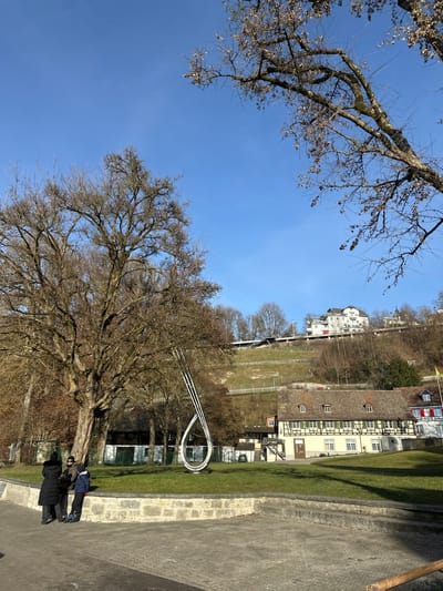 The image captures an outdoor scene in Neuhausen am Rheinfall, Switzerland, under a clear blue sky. In the foreground, a paved area transitions to a low, grey stone retaining wall. Three individuals are located on the paved surface near the wall. One individual wears a dark coat and hat, standing with their back partially to the viewer. Adjacent to them, another individual in a dark jacket and hat also stands. A third, shorter individual, dressed in a blue jacket and dark pants, sits on the stone wall, facing the other two.
The mid-ground features a large deciduous tree with bare branches to the left. To its right, centered on a manicured grassy area, is a prominent modern sculpture, possibly Roman Signer's "Tropfen" (Drop). The sculpture is constructed from multiple parallel, light-colored or white metal tubes, forming an elongated, vertical loop or teardrop shape. Behind the grassy area, a traditional building with a brown multi-gabled roof, cream-colored walls, and green-shuttered windows is visible.
The background reveals a steep, terraced hillside. Sparse vegetation, primarily bare trees and grass, covers the incline. An elevated dark structure, consistent with a railway line or bridge, crosses the hillside horizontally. Further up the slope, a multi-story white building with a red roof is partially obscured by trees. The bare branches of the trees indicate either late winter or early spring conditions, under bright daylight.