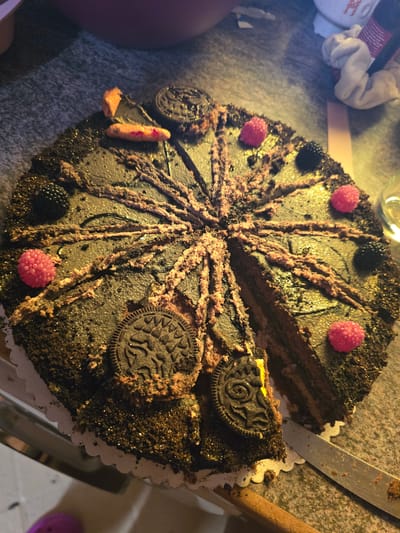 A partially sliced chocolate cake sits on a white paper plate, decorated with pink frosting lines radiating outwards, Oreo cookies, blackberries, and pink gumdrop candies. A slice has been removed, and a silver knife rests nearby. The cake is positioned on a surface near a white cloth, a wooden stick, and a glass jar. The photo was likely taken indoors. While the image does not indicate the location is Zirndorf, Germany, the picture's contents do not contradict such a setting.