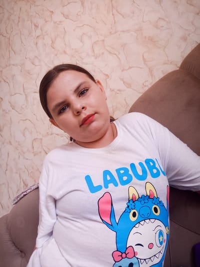 This media file is a portrait of a young woman indoors, likely in a home environment, as suggested by the patterned wallpaper and upholstered furniture. She is seated and leaning back, looking towards the camera with a neutral to slightly serious expression. Her hair is pulled back, and she appears to be wearing makeup, including lipstick.
She is wearing a white long-sleeved t-shirt with a cartoon character graphic. The character is blue with large eyes and ears, and is wearing a hat adorned with flowers. The text "LABUB" is visible in large blue letters above the character.
The lighting in the image is soft and appears to be artificial, as there are no strong shadows suggesting natural daylight. The background consists of a textured, light-colored wall with a marble-like pattern and the armrest of a brown sofa. The overall atmosphere is relaxed and casual. No specific time of day or weather conditions can be determined. The location context is Mazyr, Belarus.