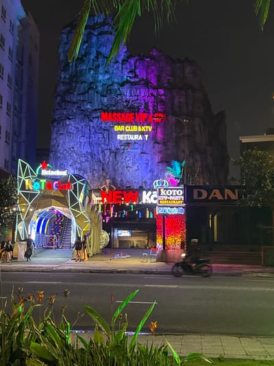 The image captures a nighttime urban street scene in An Hải Ward, Vietnam. A multi-story commercial building, centrally located, features a facade designed to mimic a rock formation, illuminated with varying shades of blue and purple light. On this facade, red neon signs display "MASSAGE VIP & SPA," "BAR CLUB & KTV," and "RESTAURANT," along with a prominent Heineken star logo.

To the left of the main building, a geometric arched entrance, bearing a Heineken star and "Koto Club" in neon, leads into the establishment with a visible, illuminated escalator. Several individuals, primarily women in dark clothing, are gathered on the sidewalk near this entrance, with more figures visible inside. A small white table with a dark top stands on the sidewalk.

Further right, another section of the building features a "NEW KOTO" neon sign and an entrance distinguished by vertical strips of multi-colored lights. A vertical sign next to this entrance reads "KOTO KTV - PARTY RESTAURANT" and "254 Vo Nguyen Giap." Two white plastic stools are positioned on the sidewalk here. To the far right, a darker building section displays partially visible letters "DAN."

On the far left, a multi-story building with multiple windows is visible against the dark sky. A two-lane asphalt road, appearing reflective, occupies the mid-ground. A blurred black scooter, with a single rider, is captured in motion on this road, moving leftward. The immediate foreground contains green foliage and plants, while palm fronds are partially visible at the top of the frame. The scene is illuminated by numerous commercial neon and LED lights, contrasting with the dark night environment.