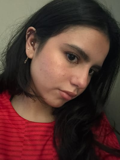 A close-up, angled selfie shows a woman with dark hair, likely in her late teens or early twenties. Her hair is long and loosely styled, falling around her face and shoulder. She is wearing a red shirt with subtle vertical stripes. Her skin tone is fair, with some freckles visible on her cheeks. She has dark eyes and eyebrows. She is wearing multiple earrings in her left ear. The background is slightly out of focus, with what appears to be a light-colored wall. The location, College Station, United States of America, is not evident from the visual information provided in the image.