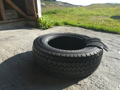 An outdoor, slightly angled, eye-level medium shot shows a car tire lying on a concrete surface in Averøy, Norway. A black glove is draped over the top of the tire. The tire, with its tread and metal studs visible, casts a shadow to its left. Behind the tire, a section of a wooden structure with exposed beams and corrugated metal is visible. Beyond that, green grass, some rocks, and a body of water under a bright sky complete the background. The scene appears to be a sunny day, with strong shadows indicating the sun's position.