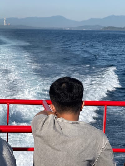 A person is seen from behind, wearing a grey t-shirt, leaning on a red railing on a boat. The boat is moving through blue water, leaving a white wake. In the distance, a hazy coastline with mountains is visible. On the far left of the distant landmass, a tall, white, industrial-looking structure with a smaller building next to it is present. The sky is clear and light blue. The overall scene suggests travel by ferry or boat. The location is identified as An Hải, Vietnam.