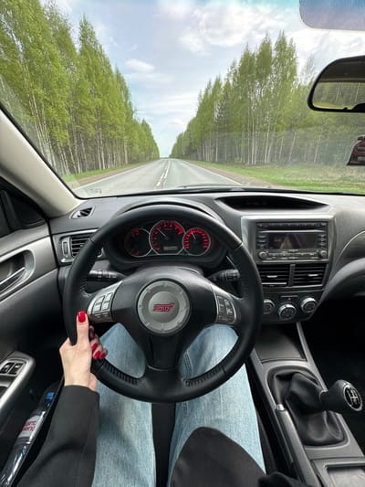 The image captures a driver's perspective inside a Subaru WRX STI, facing forward on a road with birch trees lining each side. The dashboard includes a factory head unit with a CD/AUX player, climate control knobs, and gauges showing the car has 175,980 kilometers on it. The steering wheel features the STI logo. The driver's hand, with red nail polish, is visible on the steering wheel. The gear shifter indicates a manual transmission. A glimpse of the exterior through the windshield shows a straight road with a white line. A water bottle is in the door.  The image could be in or near Kamennyi Kliuch, Russia.