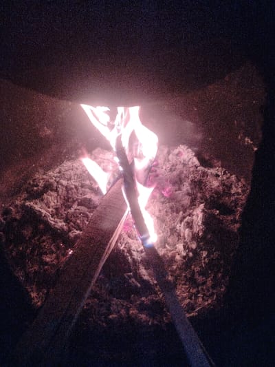 Within a dark, enclosed structure in Nurli zamin, Uzbekistan, a fire is actively burning. Multiple wooden pieces, including at least one plank, serve as fuel. Bright orange, yellow, and red flames, with localized blue coloration at their bases, emanate from the wood. A substantial bed of glowing embers and white-grey ash surrounds the combustion, indicating ongoing burning and residual material. The scene is illuminated solely by the fire, contrasting with the dark interior of the containment structure and the indistinct background. The upper edge of the dark structure is visible across the top portion of the frame. No individuals or specific interactions are depicted.
