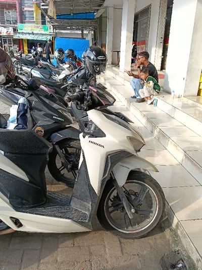 This image captures a street scene in Sumberejo, Indonesia, under bright daylight. In the foreground and mid-ground, a dense row of numerous scooters, predominantly black and white Honda models, are parked on a paved surface. Multiple motorcycle helmets, including a prominent black full-face helmet with a clear visor, are visible on or near the parked vehicles. On the immediate left, a white Honda scooter is partially visible.

To the right, two individuals are seated on the lower concrete steps of a building. An adult male with short, dark, curly hair and a beard, wearing a black t-shirt and blue jeans, sits holding a mobile phone in his left hand. Next to him, on a lower step, is a young child wearing a green and white jacket and light-colored pants, looking towards the left foreground. Scattered refuse, possibly plastic packaging, is visible on the steps near the child.

In the background, behind the parked motorcycles, commercial buildings are visible, including one with a green sign reading "NAFIS SHOES." A blue tarp or fabric structure is also visible further back. Several indistinct figures can be observed near the buildings and other motorcycles in the background. Shadows cast by the vehicles and buildings indicate strong sunlight.
