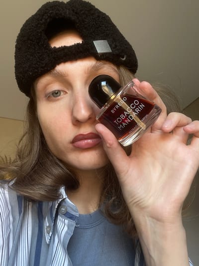 A woman wearing a black beret and a striped blue and white button-down shirt is holding a bottle of BYREDO Tobacco Mandarin Extrait de Parfum in front of her face. The bottle is held in her right hand, with the cap covering her right eye. Her lips appear to have a glossy, wine-colored lip product on them. Her hair is light brown and wavy. The background is a light beige wall. There is no evidence to suggest the location is in Sepych, Russia.