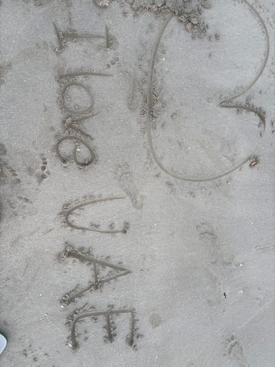 The image shows the words "I love UAE" written in the sand on a beach. The writing is in large, blocky letters and appears to be freshly made. A heart shape is also drawn next to the words. The sand is light gray and appears to be damp, with some small shells and pebbles scattered around. There are also a few footprints visible in the sand, indicating that someone has been walking on the beach. The image appears to have been taken from a slightly elevated angle, looking down at the sand. The scene is consistent with a beach in Ajman, United Arab Emirates, a popular destination for recreation and tourism.