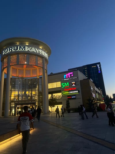 The image depicts the Forum Kayseri shopping mall and the adjacent Radisson Blu hotel at dusk. The mall's iconic circular entrance tower, illuminated with warm orange light through ornate geometric patterns, stands prominently on the left. Large glass windows reveal the interior of the mall, with shoppers visible inside. To the right, the multi-story Radisson Blu hotel displays its prominent blue signage against the darkening sky.
Several signs advertising shops and services within the mall are visible, including "FORUM KAYSERI," "5M MIGROS," "Media Markt," "Paribu Cineverse," "Boyner," and "Koton." The overall scene suggests a vibrant urban environment as the evening progresses, with people walking on the paved plaza in front of the mall. The sky transitions from a deep blue overhead to a lighter hue near the horizon, indicating the fading light of day. The ground is paved with dark tiles, and a textured tactile paving strip runs along a walkway.