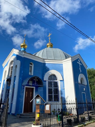 The image shows a bright blue and white church with golden domes. The church has arched windows and decorative elements. In front of the church, there is a black metal fence. The sky is blue with white clouds. The image appears to be taken on a sunny day. Based on the architectural style and the presence of Orthodox Christian symbols, this is likely an Orthodox church. Without further information, it is not possible to confirm the specific location of the church in Kazan, Russia.