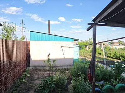 In Tashla, Russia, on a sunny day, a backyard scene is captured. The foreground features garden beds with various green plants and some small pink flowers. To the left, a brown, vertical-slatted fence runs along a brick wall. A light-colored building with a turquoise corrugated metal roof is prominent in the middle ground. A rusty metal chimney extends from the roof. To the right, a makeshift wooden structure with a dark, angled roof provides shade. A towel hangs on a wooden post in front of the structure. In the background, a densely vegetated garden area is visible, with some small houses and a utility pole with wires in the distance under a blue sky with scattered white clouds.