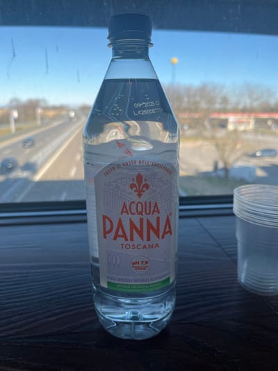 This image features a plastic bottle of Acqua Panna natural mineral water, positioned indoors on a dark surface next to a window. The bottle is the main subject, and behind it, a clear plastic cup or container is also visible, out of focus. The scene is set in Dorno, Italy, looking out onto a daytime view through the window.
The background, seen through the window, shows an out-of-focus outdoor environment with bright sunlight, suggesting clear weather. A road or street is visible, with hints of vehicles, trees, and buildings further in the distance. The lighting indicates it is daytime.
Notable text on the water bottle includes:
- A date "09- 2025 22" and a lot number "L4250087704", likely indicating an expiration date and production code.
- The brand name "ACQUA PANNA TOSCANA" (Acqua Panna from Tuscany).
- A slogan "TUTTO IL GUSTO DELL'EQUILIBRIO" which translates to "All the taste of balance".
- Information about the water: "pH 7,9 NATURALE ACQUA MINERALE NATURALE OLIGOMINERALE" (pH 7.9 Natural Oligomineral Natural Mineral Water).
- An environmental note: "BOTTIGLIA CON PLASTICA RICICLAIR", indicating the bottle is made with recycled plastic.