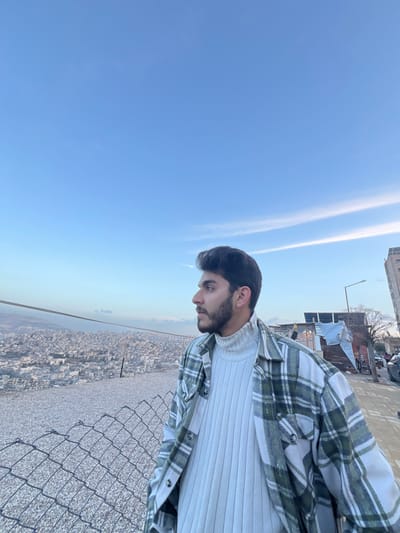 A young man stands on a rooftop overlooking a dense urban landscape stretching towards the horizon. He wears a white ribbed turtleneck sweater and a green and white plaid flannel shirt. A chain-link fence lines the edge of the rooftop, offering a safety barrier. The sky is a clear, bright blue with faint cloud streaks. The scene suggests a high vantage point, possibly a building or a viewpoint in a city. The photo is taken from a slightly tilted angle.