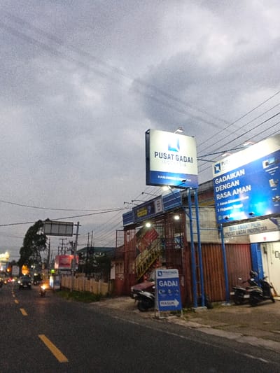 This image captures an urban street scene at dusk or dawn under an overcast, grey sky in Phước Hòa, Vietnam. A two-lane asphalt road, marked with a double solid yellow line and a broken yellow line, occupies the lower left. On the right side of the road, a commercial building features prominent blue and white illuminated signage. The largest sign, elevated on poles, reads "PUSAT GADAI INDONESIA" (Indonesia Pawn Center) with a square logo. Below and to its right, another large sign displays "GADAIIKAN DENGAN RASA AMAN" (Pawn with peace of mind), providing contact details. A smaller, ground-level blue and white directional sign indicates "GADAI" (Pawn) with an arrow and "MASUK" (Enter). The building structure visible includes a reddish-brown, possibly metal, exterior with a visible staircase on the left side of the building's facade. Several motorbikes are parked on the paved area in front of the building. Overhead utility wires crisscross the frame above the street and building. On the left side of the road, distant vehicle headlights, likely from motorbikes, are visible, and a large, unlit billboard structure stands further down the street. Trees and other distant buildings complete the background.