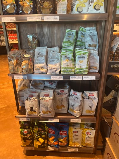 The image displays an indoor scene, likely within a grocery store or specialty food shop in Dorno, Italy, given the context. The primary subjects are various packaged food items, predominantly snacks and crackers, arranged on brown wooden shelves with clear plastic dividers.
On the left side of the shelving unit, there are bags of "TARALLINI" in green and white packaging, some labeled "WHOLEGRAIN" or "ONE SEED." Below these are white bags of "TARALLI ARTIGIANALI," including a variety labeled "EXTRA VERGINE OLIVA." Further down and to the left, there are clear packages of "MINI" branded crackers or snacks. Adjacent to these, on the far left, are larger white and yellow bags of potato chips, possibly featuring truffle or other flavors, some with prices like "4.00 EUR" or "3.90 EUR".
The right side of the display features several brands of potato chips. The top shelves hold white bags of "AMICA Alfredo's Salt" chips, recognizable by the salt shaker graphic. Below these are "AMICA Alfredo's Ruffle" and "AMICA Alfredo's Truffle" chips, distinguished by their respective flavor icons. Further to the right, yellow bags of "PATA spesse" chips are visible, followed by blue bags of "AMICA Chipi" or "AMICA Chips ENERGY." The bottom right shelves contain dark green/black bags of "PATA" brand chips.
Many products have visible price tags, such as "3.90 EUR," "3.00 EUR," "2.80 EUR," and "2.70 EUR," indicating that items are for sale. In the background, behind the shelves, there are dark wooden elements and some cardboard boxes, one with the partial text "BAB" and another "EA." The lighting suggests an indoor setting, and no people or specific activity are visible, only the neatly organized merchandise. The prominent Italian text on many packages further reinforces the location context.