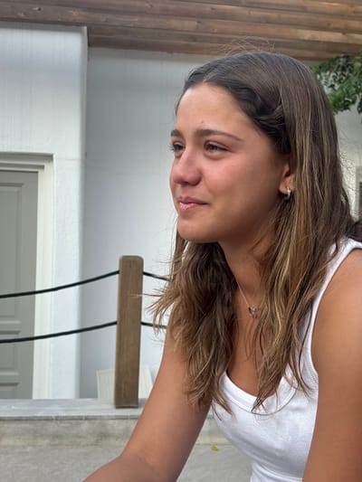 A young female subject is depicted in an outdoor setting in Platys Gialos, Greece. She is shown in profile, facing to the viewer's left. Her long, light brown hair, streaked with lighter highlights, cascades over her shoulders. She wears a white ribbed tank top, a silver necklace with a small heart-shaped pendant, and small silver hoop earrings on her visible left ear. Her complexion appears tanned. The background consists of a white stucco building with a light grey panel door visible to the left. A wooden support post with a black rope barrier extends horizontally behind the subject, positioned above a grey stone or concrete platform. Overhead, exposed wooden beams suggest a covered outdoor structure. Minimal green foliage is present in the upper right background. The scene is illuminated by natural daylight.