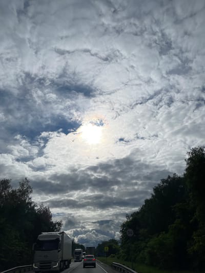 The image shows a view looking upwards and slightly to the right, capturing a sky filled with textured, layered clouds, partially obscuring the sun. The scene includes a road with vehicles, including a white truck, and trees lining the roadside. A road sign with a '50' speed limit is visible. This image was not taken in the city of Ol'khovka, Russia.