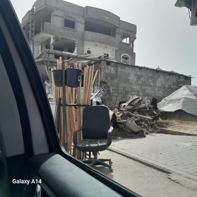 The image shows a view from inside a vehicle, possibly a car, looking out at a building in the city of Nuseirat in the Palestinian Territories. The building appears to be heavily damaged, possibly due to conflict or demolition, with visible structural damage, missing walls, and signs of decay. In the foreground, there is an office chair and construction materials, including lumber and black buckets, stacked on some kind of shelving unit. The scene suggests the aftermath of destruction and potential ongoing construction or repair efforts. The sky is overcast. The phone is a Galaxy A14.