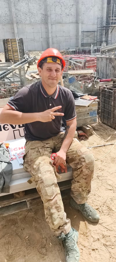 A man in a hard hat and camouflage pants is sitting on a white surface outdoors at a construction site. He is wearing a dark polo shirt with red and white trim. His right hand is raised, showing a peace sign. In his left hand, he is holding a pair of red-handled pliers. He has a tattoo on his left forearm. The background shows a large concrete wall, scaffolding, and construction materials. The ground is covered in dirt and sand. The image appears to be taken in Primorye, Russia, a region known for its industrial and construction activities.
