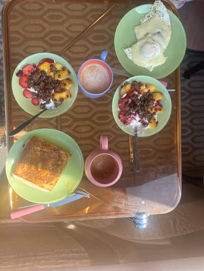 The image depicts a breakfast meal served on a glass tray. The tray sits indoors, likely near a window, suggested by the shadows cast on the right side. The breakfast consists of four plates and two mugs. A green plate at the top left contains two fried eggs covered in cheese. Beside it is another green plate holding a yogurt bowl with peaches, strawberries, and granola. Below the egg plate is a blue mug containing a beige liquid. Next to it is a similar bowl to the first, filled with yogurt, peaches, strawberries, and granola. To the right is a pink mug filled with dark liquid. A green plate with a piece of French toast, with a pink-handled knife and fork. The location of the image is unknown and there are no people in the image.