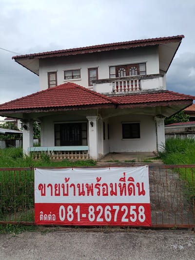 This is a daytime, eye-level, outdoor photograph of a two-story house in Ban Patan, Thailand. The house has a white facade with a red tiled roof. A white banner with red text is attached to a rusty metal fence in front of the house. The text on the banner reads "ขายบ้านพร้อมที่ดิน" (meaning "House for sale with land") and "ติดต่อ 081-8267258" (meaning "Contact 081-8267258"). The house appears to be unoccupied, with overgrown grass in the yard. The sky is overcast.