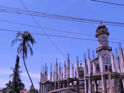 The image shows a building under construction in Keudealue Ie Puteh, Indonesia. The structure appears to be an expansion or renovation of an existing mosque. The concrete framework of the new construction surrounds the older, completed sections of the mosque, including a minaret and a dome. Reinforced steel bars protrude from the tops of the concrete columns. Overhead, a network of power lines crisscrosses the sky. A tall palm tree stands to the left of the building. Another shorter palm tree is next to the structure. The sky is blue and clear.