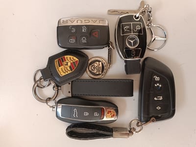 The image displays a collection of car keys and keychains arranged on a light-colored, flat surface. The keys include those for Jaguar, Mercedes-Benz, Porsche, and BMW vehicles, identified by their respective logos and symbols. Keychains include a Porsche emblem, a sparkling keychain with a skull motif, a black leather strap, and a Mercedes-Benz emblem. The lighting suggests an indoor setting, possibly a desk or table. No people are visible in the image. The location in Mohyliv-Podilskyi, Ukraine, cannot be determined from the visual information provided.