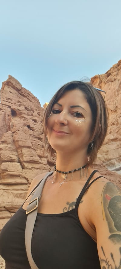 A selfie captures a woman in the city of Hevel Eilot Regional Council, Israel. She has fair skin, brown hair, and is wearing glitter on her face. Her left shoulder is visible, revealing a tattoo, and she is wearing a black tank top and a necklace with silver chains. A strap is across her chest. The background shows a blue sky and a large rock formation.