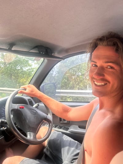 A smiling, fair-skinned man with blonde hair drives a car. He's wearing a grey tank top and dark shorts. His right hand is on the steering wheel, which has a Mercedes logo. Sunglasses are clipped to the car's visor. The car is moving; trees are visible through the front window. The photo is taken from an unusual angle, with the camera tilted to the side.