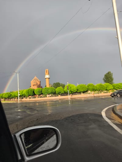 This is a medium shot of a street scene in Artashat, Armenia, taken from inside a vehicle. The foreground features a car's side mirror, reflecting the interior and the rainy street. The road is wet and shows signs of wear, with a white lane marker curving to the right. In the midground, a row of neatly trimmed, vibrant green trees lines a decorative fence. Beyond the trees stands a red brick religious building with a domed roof and a tall, slender tower. A faint double rainbow arcs across the dark, overcast sky, indicating recent rain. The sky is a gloomy grey, with some lighter patches suggesting the sun may be attempting to break through. Power lines are visible crisscrossing the sky. A street lamp with traffic lights stands on the left, and a black car drives away in the distance on the right. The overall mood is somber due to the weather, but the rainbow offers a touch of hope. There is no visible text in the image.