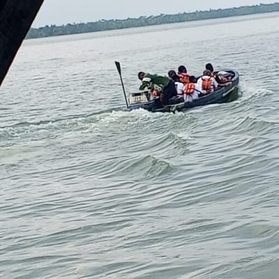 An open, motor-powered boat traverses a waterway in Koko, Nigeria, under an overcast sky. The dark-colored vessel creates a wake, indicating forward motion. Approximately 7-8 individuals are visible within the boat; at least four are wearing orange life jackets. One person in the bow area wears a green garment, while another in the stern, possibly operating the boat, wears a dark top. A paddle stands vertically near the stern, adjacent to a white box-like object marked "JIS". The water surface is choppy with discernible waves. A distant treeline defines the horizon, indicating land. A dark, out-of-focus object partially obscures the upper left foreground.