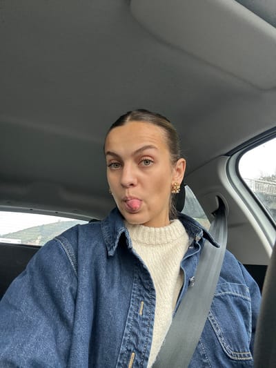 A young adult female is depicted inside a vehicle, reportedly located in Saint-Cyr-sur-le-Rhône, France. She is positioned centrally in a close-up, frontal portrait. Her light brown hair is pulled back, and her green-brown eyes are directed at the camera as she extends her tongue. She wears ornate, gold-toned stud earrings in both ears. Her attire consists of a cream-colored, ribbed knit sweater underneath a dark blue denim shirt or jacket. A dark gray seatbelt is fastened across her left shoulder and chest. The vehicle's interior features a gray headliner and window trim. Through the rear left window, a sloped, vegetated hillside is visible under an overcast sky. Through the right window, a gray concrete or stone structure, possibly a bridge railing, is partially visible, also under an overcast sky. No specific geographical landmarks within Saint-Cyr-sur-le-Rhône are discernible.