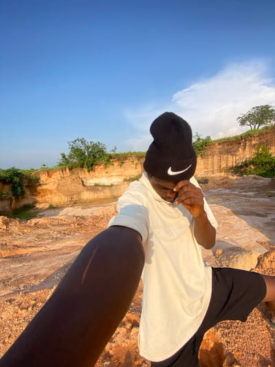 In Ufu Esioni, Nigeria, a person wearing a black Nike beanie and a cream-colored t-shirt stands in a quarry. The person is holding their right hand to their face. The ground is a mix of dirt and rocks, with layered sediment walls visible in the background. Green foliage grows on top of the quarry walls and in scattered patches around the site. The sky above is clear blue with a few wispy clouds. The sun casts a warm light on the scene, creating shadows.