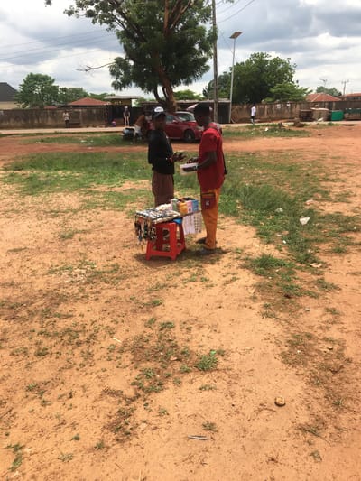 This image, taken in Isieke, Nigeria, captures an outdoor scene featuring two individuals engaged in what appears to be a roadside transaction under an overcast sky. The ground is largely reddish-brown dirt with sparse patches of green grass and some scattered litter.
On the right, a person in a red long-sleeved top and light brown trousers stands behind a small stall. The stall is formed by a red plastic stool topped with a wooden box or tray, displaying an assortment of small items such as sunglasses, keychains, or jewelry. A small pouch on the person's hip features a faint "SS" or "55" logo. Facing this vendor is another individual, dressed in a dark top and trousers, wearing a helmet, who seems to be a customer examining the merchandise.
In the background, a red car is partially visible behind the vendor. Further back, on the left, a large tree dominates the mid-ground. Beyond the tree, several modest buildings with reddish roofs can be seen under a cloudy, grey sky. Utility poles and power lines crisscross the upper left of the frame. The overall impression is a casual, everyday setting, likely a local market or roadside selling point during the day. No clearly legible text is visible other than the faint logo on the vendor's pouch.