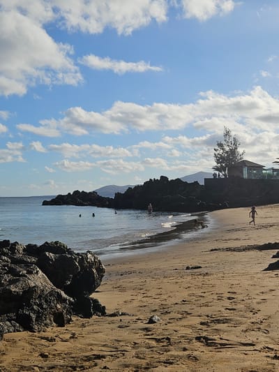 The image depicts a sandy beach with dark volcanic rocks jutting out into the clear blue water under a partly cloudy sky. In the shallow water, several individuals are visible swimming or wading. On the sandy shore, a young child in a striped swimsuit runs towards the right side of the frame, while another child kneels and plays in the sand. Further back, a building with a brown roof and large windows is situated on a rocky outcrop, with a few people standing on a balcony or terrace. A solitary tree with sparse foliage stands near the building. The overall environment suggests a sunny day at a coastal location, likely in Tías, Spain, known for its volcanic landscapes and beaches.