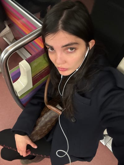 A young adult female is depicted in a medium close-up, seated inside a public transport carriage, characteristic of the Paris Metro, within the city of Saint-Mandé, France. She possesses dark hair, centrally parted, and brown eyes, looking directly at the camera with a slight pout. White wired earphones are inserted into both ears, their cord visible descending over her dark, buttoned coat. A brown leather shoulder bag, bearing a repeating monogram pattern, is slung across her body. Her left hand rests on her lap, partially obscuring a dark object with red detailing. The seat to her left features a multi-striped textile pattern in shades of purple, green, orange, and pink. A silver-colored metal grab bar is present adjacent to the seat. The visible floor surface is a reddish-pink color.