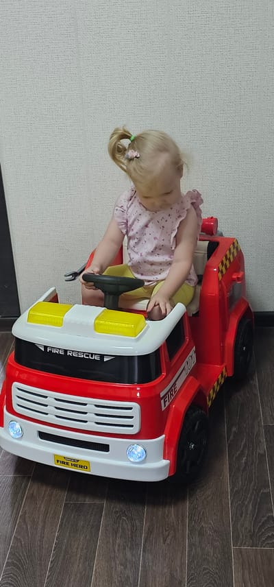 A young child with blonde pigtails, adorned with a small pink bow in her hair, is seated in a red toy fire truck. She wears a pink shirt with a floral pattern and yellow pants. The child is actively engaged with the toy, her hands on the steering wheel. The fire truck is bright red with white accents, featuring yellow headlights and a grill. Text visible on the truck includes "FIRE RESCUE" and "FIRE HERO." The scene appears to be indoors, with a textured light-colored wall on the left and dark wooden flooring on the right. The lighting suggests it could be daytime.