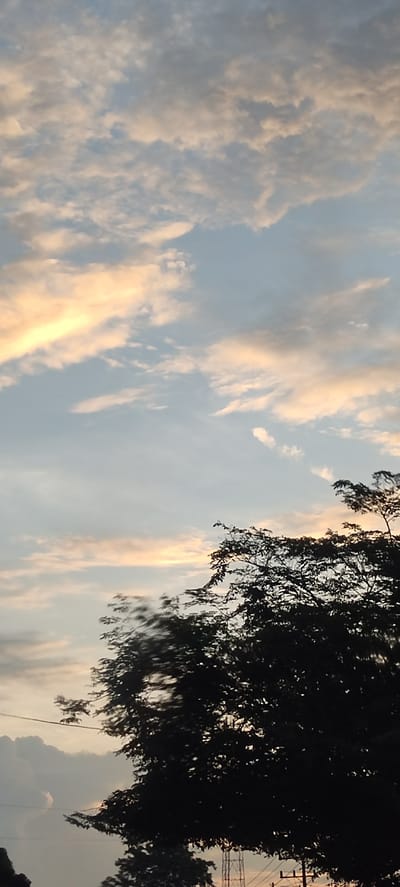 The image is taken outdoors in Aluegunto, Indonesia, depicting a partly cloudy sky at sunset or sunrise. The clouds are a mix of white, gray, and light orange hues, indicating the presence of sunlight. In the lower portion of the image, the silhouette of a tree with dense foliage is visible. Wires and an antenna structure can be discerned among the trees.