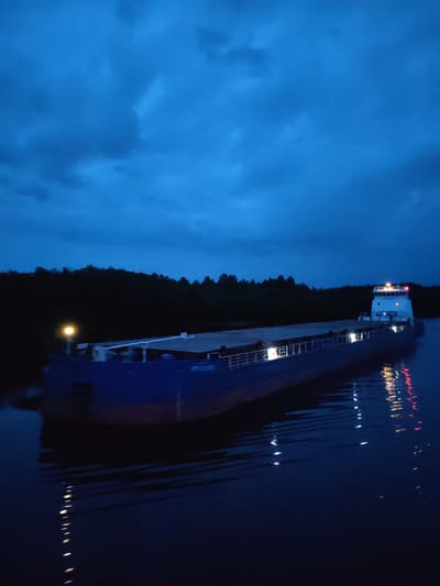 The image depicts a large, flat-bottomed barge navigating through dark, choppy waters under a deep blue, cloudy sky. The barge is illuminated by several lights, including a bright yellow light on the front and white lights along its length. The stern of the barge features a white superstructure with additional lights, some of which are red. A dark line of trees forms the background, suggesting a wooded shoreline. The reflection of the barge's lights shimmers on the water's surface. The overall scene conveys a sense of twilight or nighttime activity on a waterway, possibly a river or canal, given the calm water and the presence of a barge designed for inland transport. The location is Yalosar', Russia.