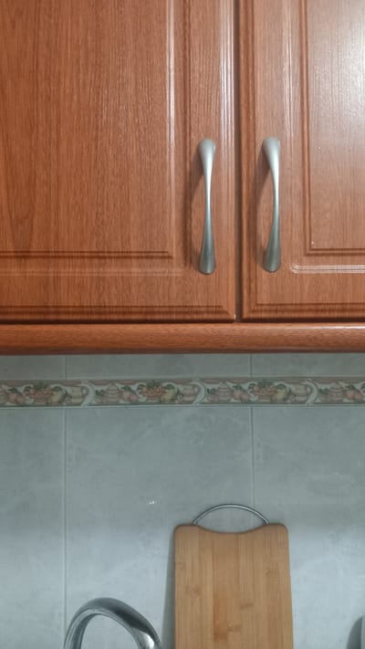 The image depicts a close-up view of a kitchen scene. At the top, there are two wooden cabinet doors with a light brown finish and vertical grooves. Each door has a silver, elongated handle. Below the cabinets, there is a tiled backsplash with a decorative border featuring an illustration of fruit baskets and pots. The lower portion of the image shows a light gray tiled wall. On the right side, a wooden cutting board with a curved top and a silver metal handle is propped against the wall. On the left, a portion of a silver kitchen faucet is visible. The overall impression is that of an interior kitchen space, likely in a residential setting. The location is identified as Oliveira do Bairro, Portugal.