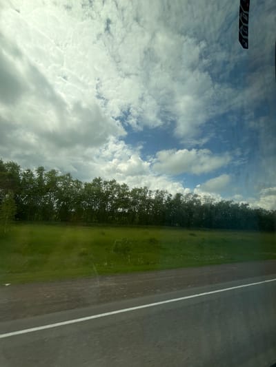 The image shows a roadside view on a cloudy day. In the foreground, a paved road with a white line is visible. Beyond the road is a grassy field, followed by a dense line of trees. The sky above is filled with a mix of white and grey clouds, with patches of blue sky peeking through. The scene appears to be an outdoor landscape, likely in a rural or semi-rural area. The image does not contain any people or specific identifiable landmarks that would pinpoint the location to Karacha-Elga, Russia.